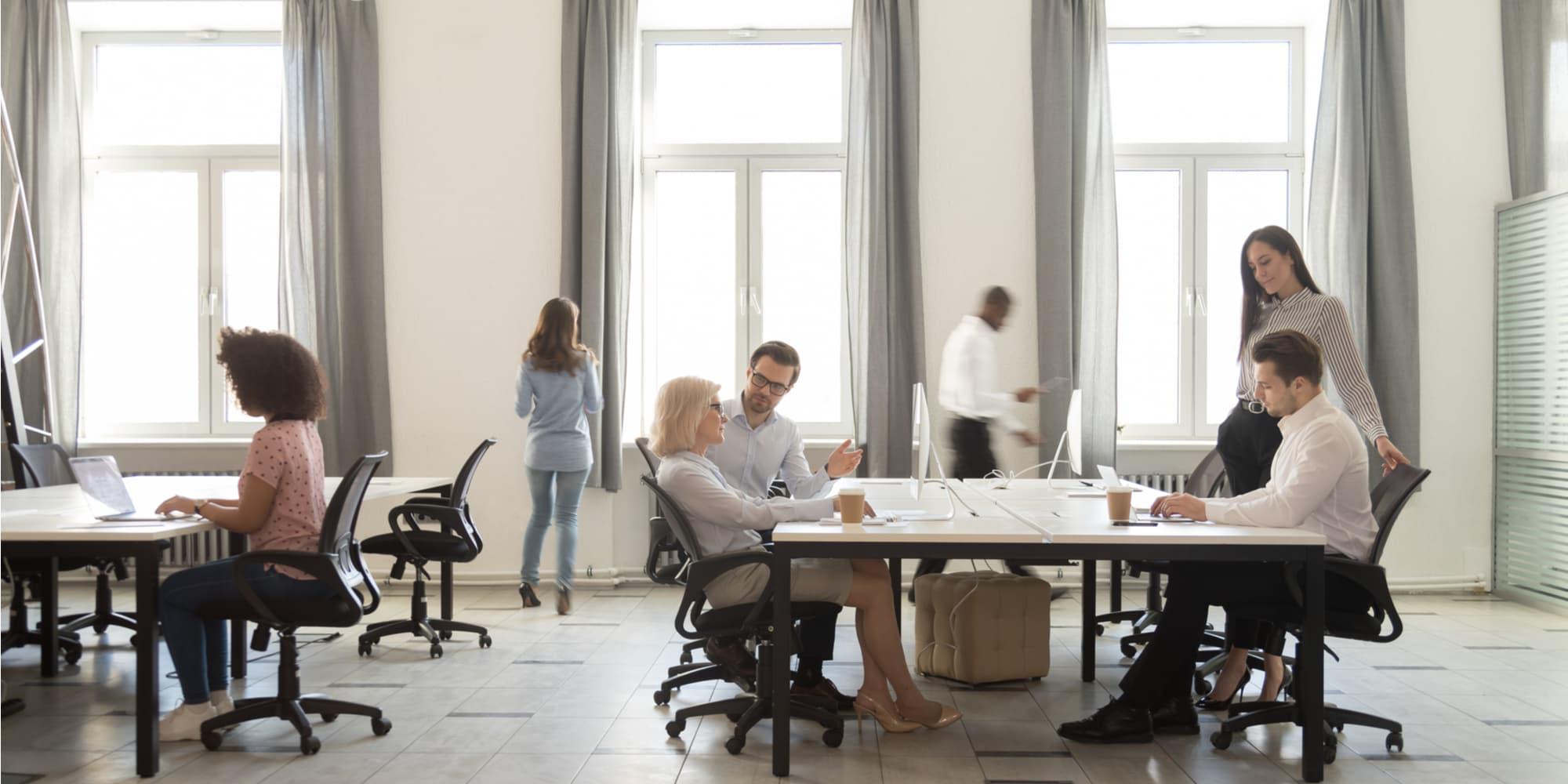 Busy company department members employees working together sitting at shared desks use computers walking in modern coworking space room interior daylight through windows